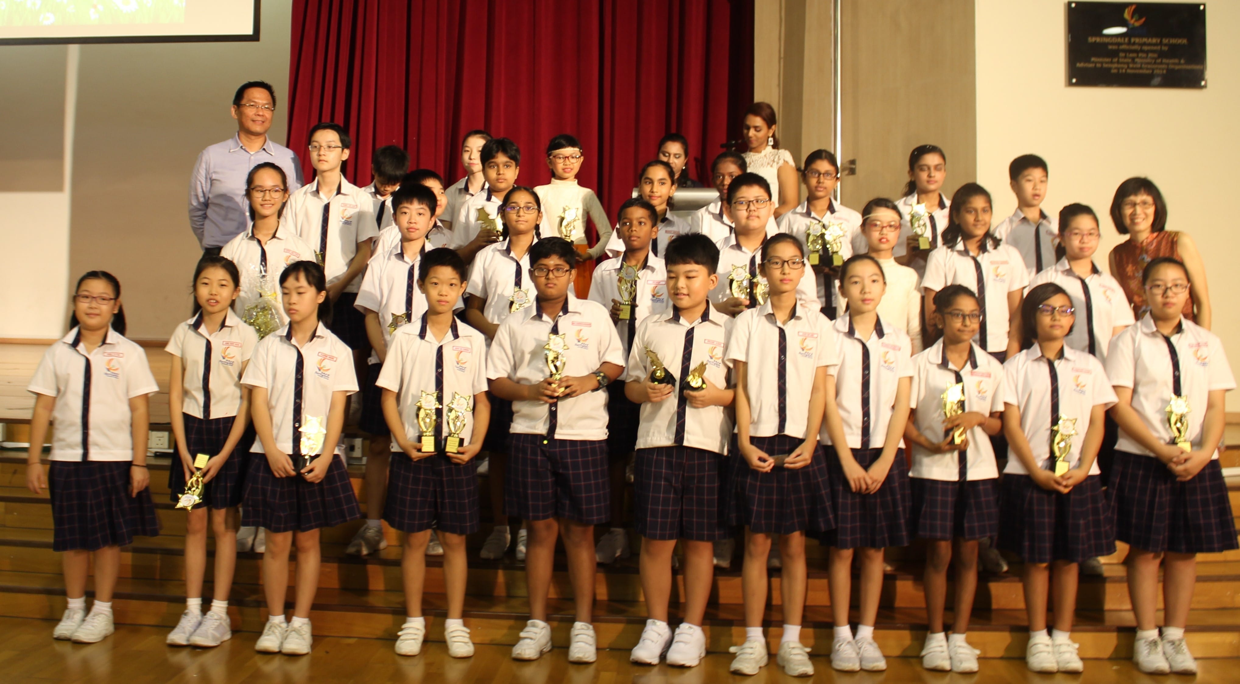 A large group of primary school students in white uniforms holding gold trophies, posing on a stage with two adults.